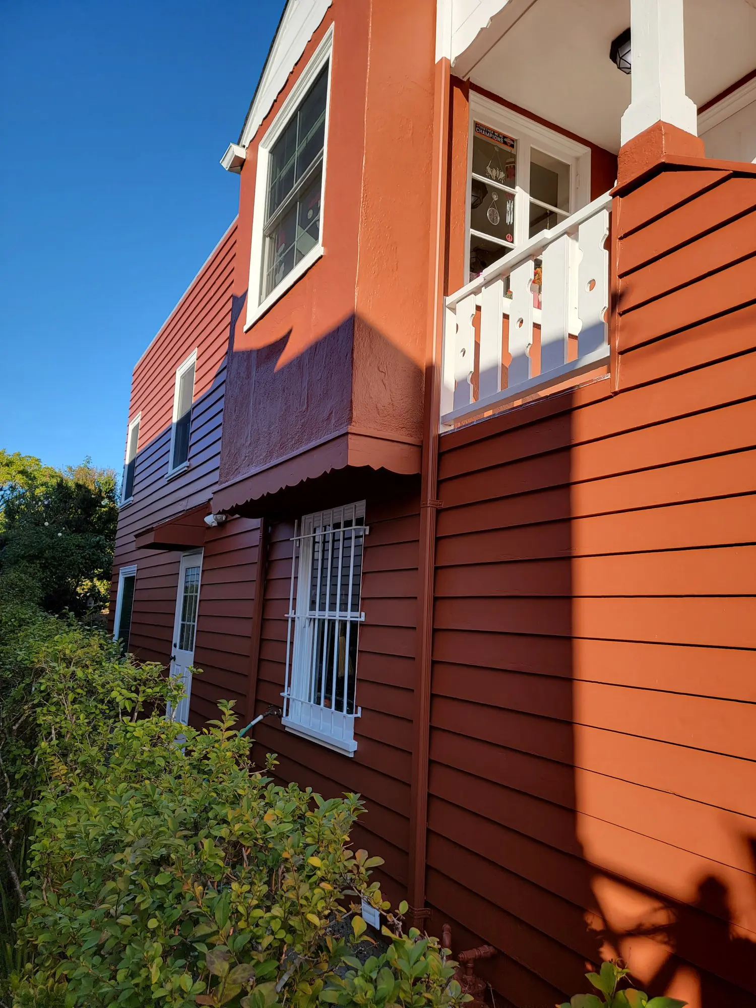 Sunlit red wooden house exterior with white window frames and green bushes.