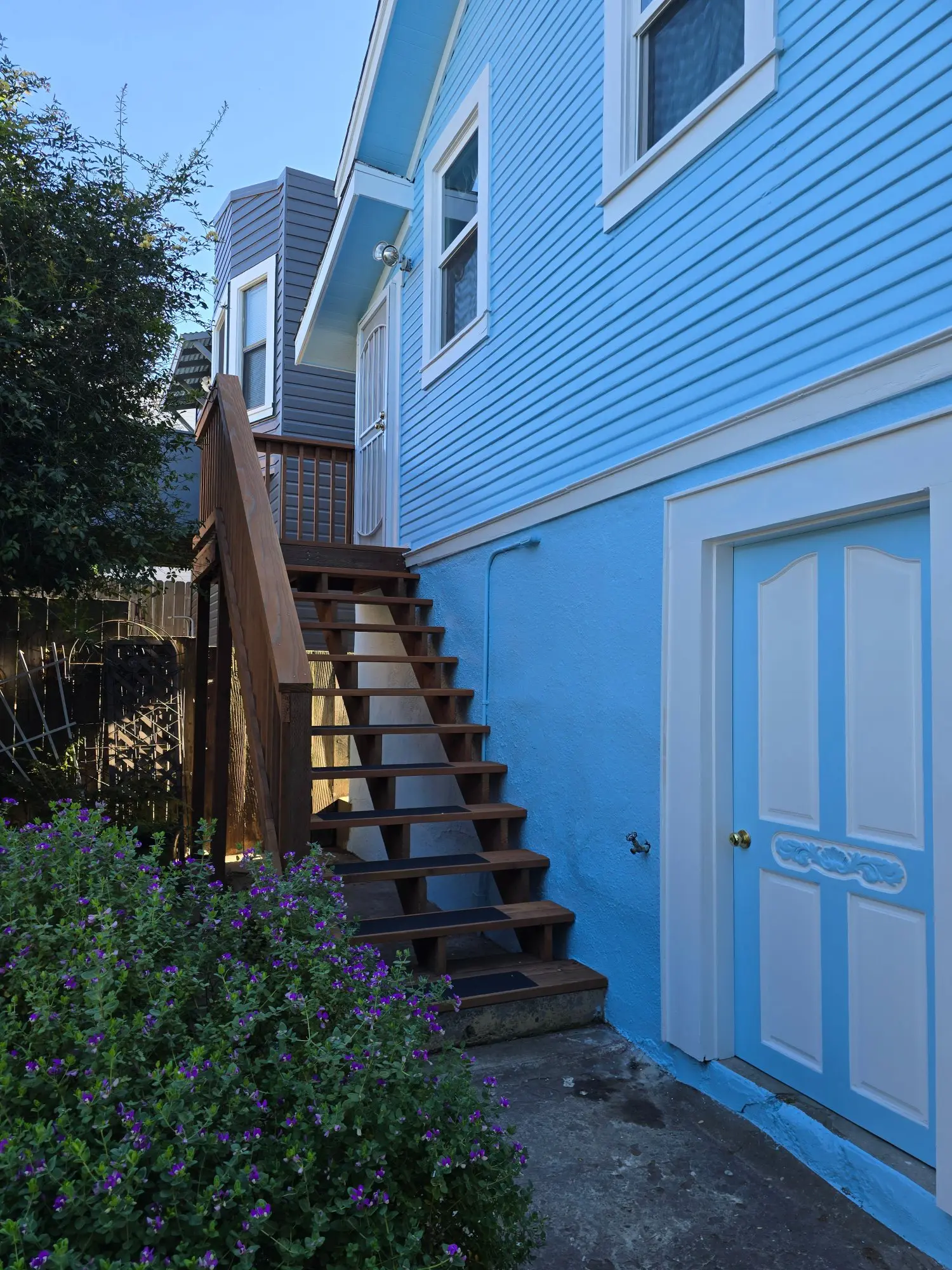 Wooden stairs lead up the side of a bright blue house with a garden nearby.