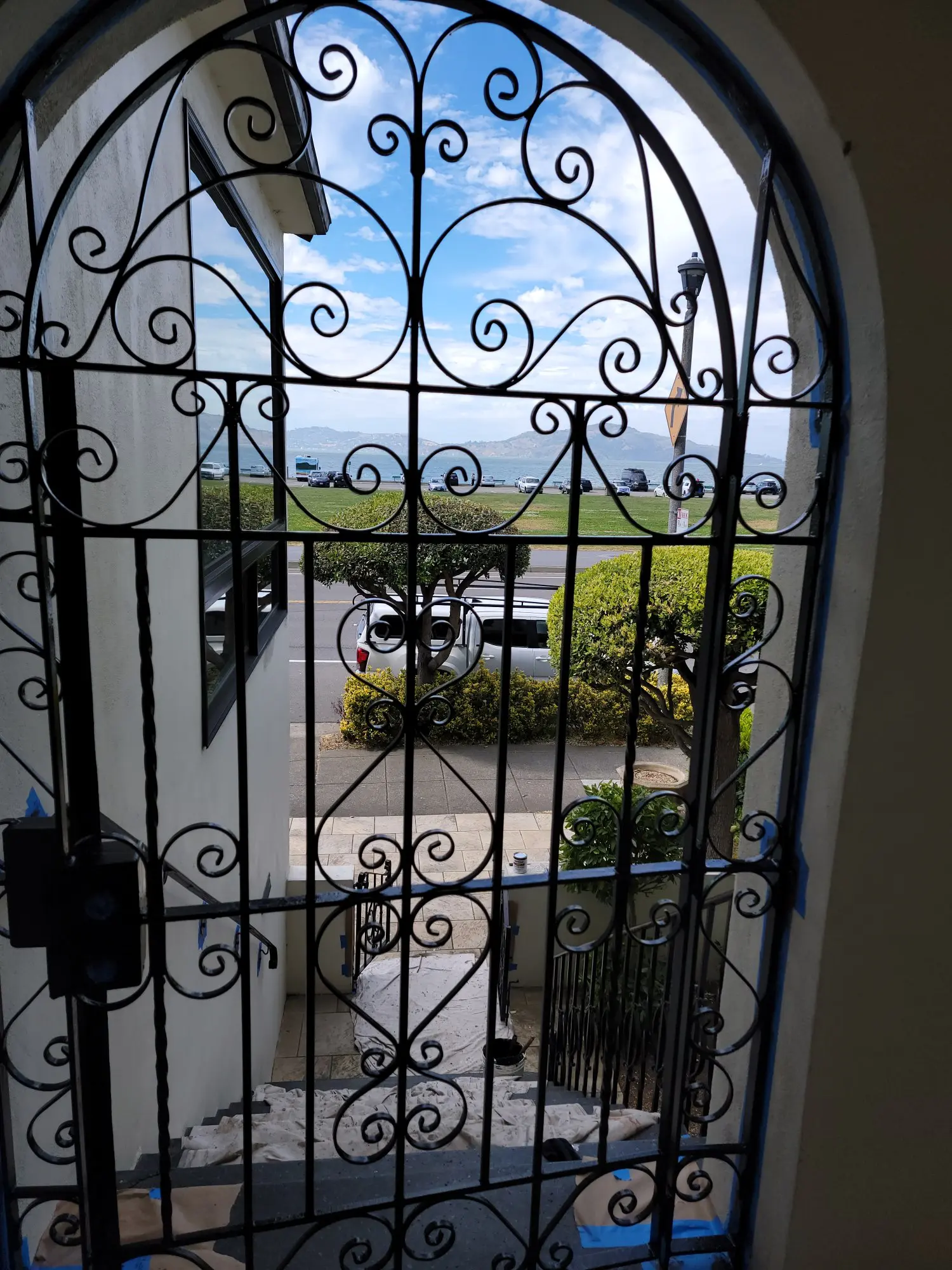 View through an ornate wrought iron gate to a sunny parking lot and greenery.
